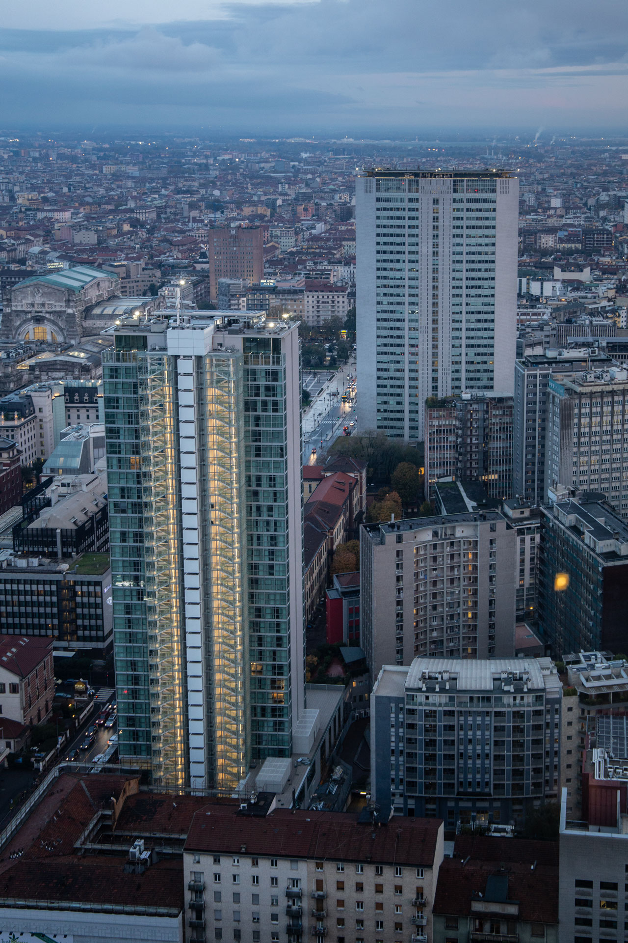 Il Pirellone al crepuscolo visto accanto a una torre residenziale illuminata, con Stazione Centrale sullo sfondo.